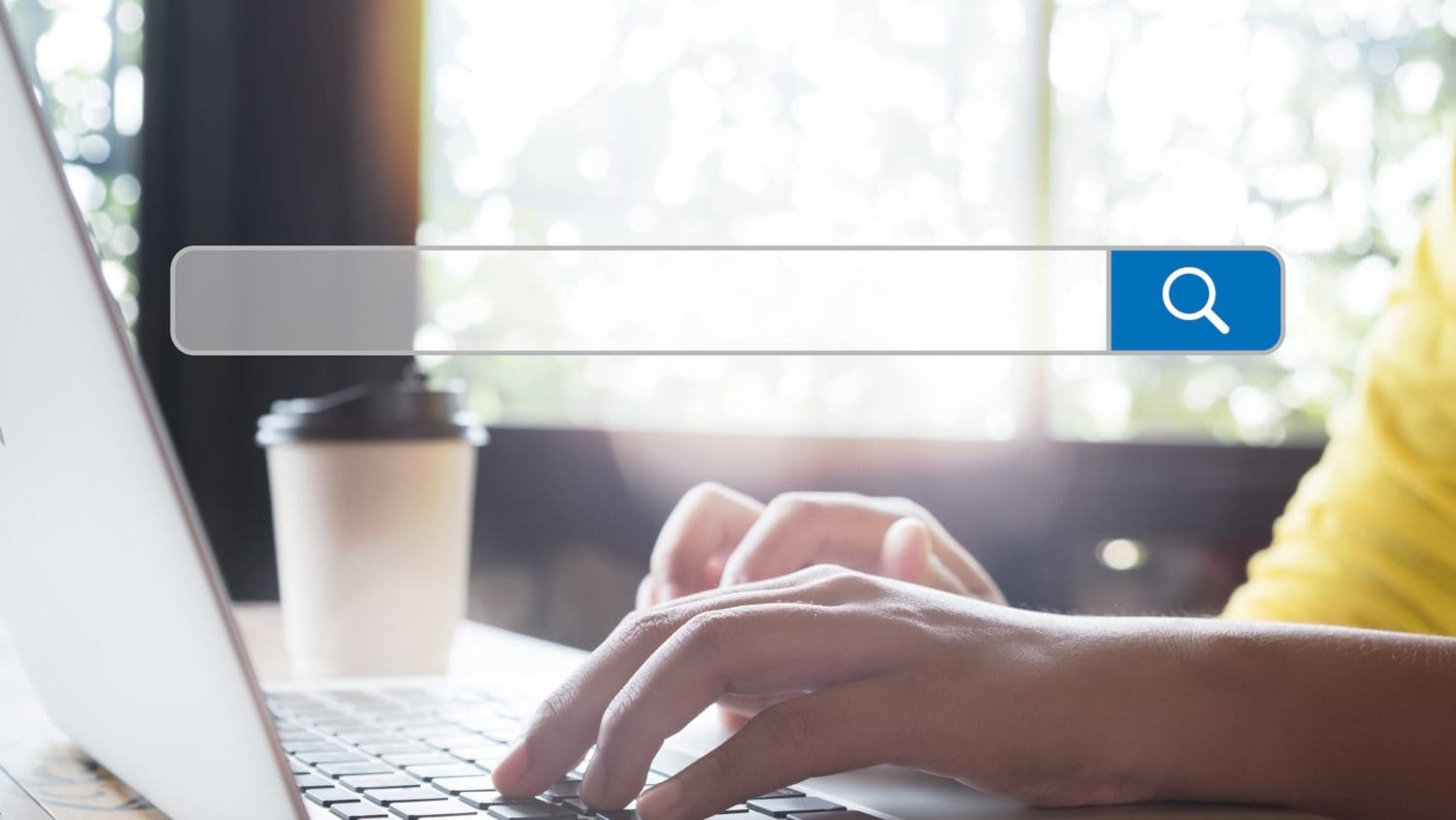 Hands typing on a laptop keyboard with a predictive search bar overlay on the image. A takeaway coffee cup is nearby, and sunlight filters through a window in the background.