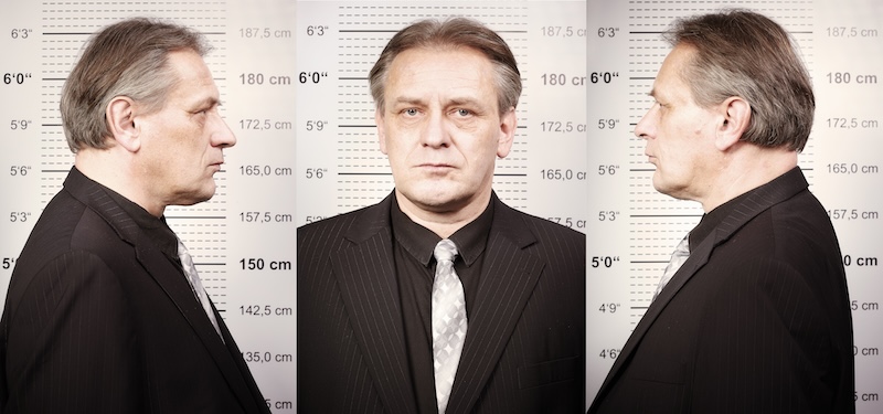 A man in a black suit with a gray tie is photographed from three angles—left profile, front, and right profile—against a height measurement background, typically used in a mugshot. With short gray hair and a neutral expression, it's important to note that are all mugshots public record.