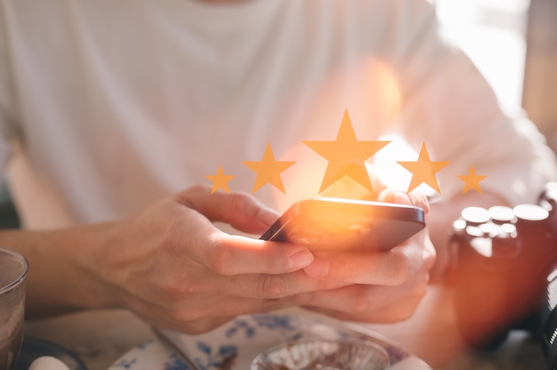 A person holds a smartphone, with a transparent overlay of five stars hovering above the device, suggesting a high rating or review. The background is softly blurred, focusing on the phone and stars.