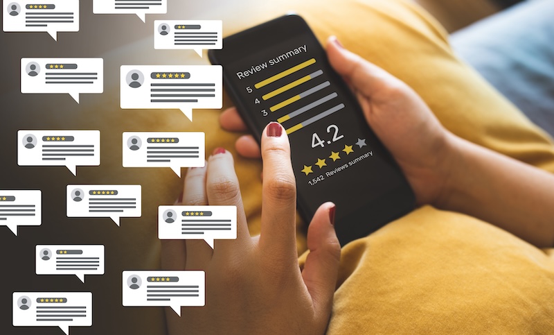 A person taps on a smartphone screen displaying a review summary with a 4.2-star rating, contemplating how to get Google reviews. Multiple speech bubbles containing comments and star ratings float on the left side of the image, indicating various user reviews. The person rests on a yellow cushion.