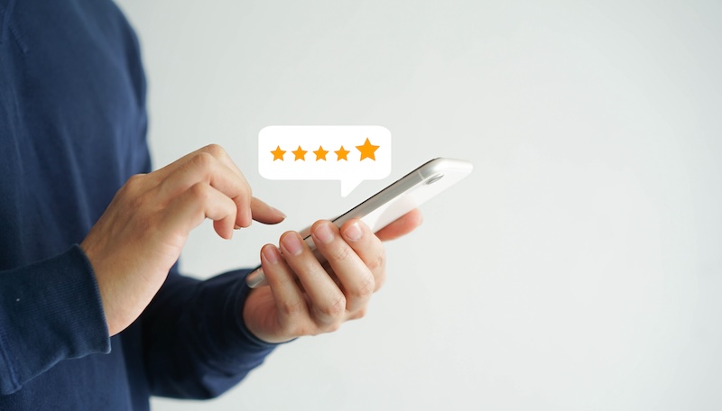 A person in a blue sweater using a smartphone displays a digital five-star rating graphic hovering above the device, symbolizing positive feedback or review.