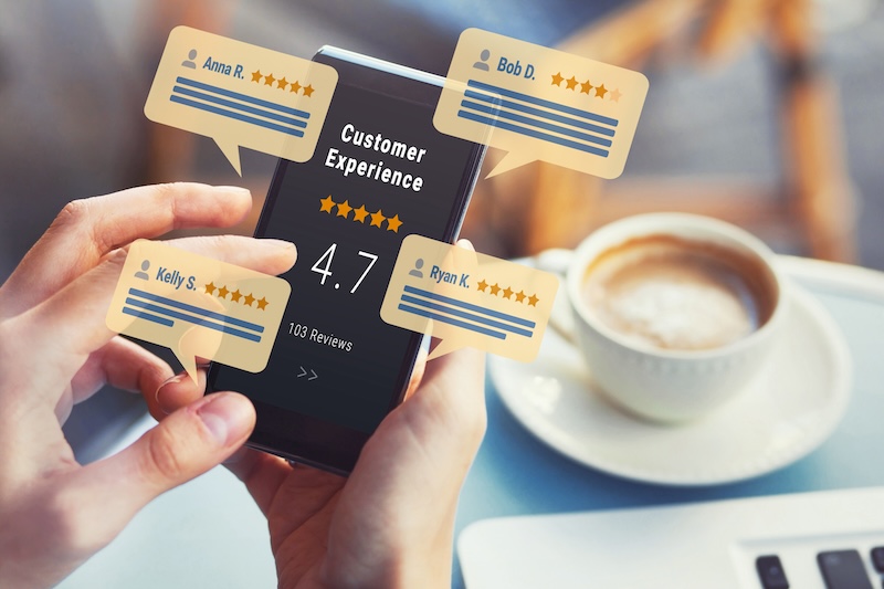 A person holding a smartphone displaying a customer experience rating of 4.7 stars with 103 reviews. Around the phone are floating review bubbles from users with five-star ratings. A cup of coffee and a saucer are on the table in the background.