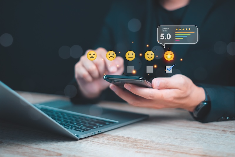 A person holds a smartphone displaying a series of emojis from sad to happy, with a "5.0" rating and stars above it. A laptop is on the desk in the background.