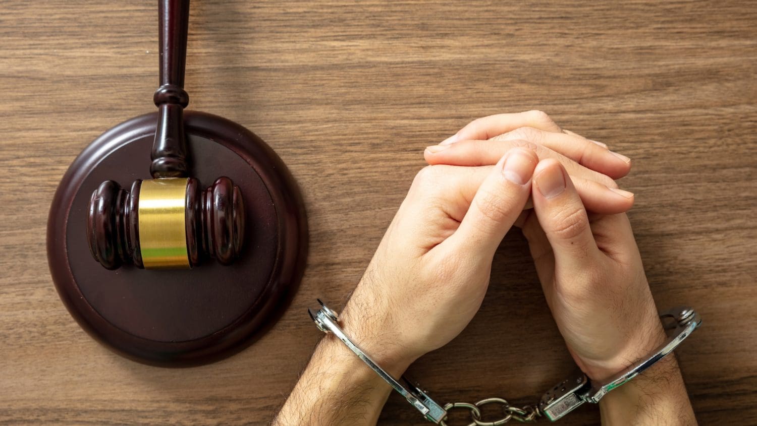 Hands in handcuffs rest on a wooden table next to a brown gavel, symbolizing the challenging journey of how to clear your record of a felony.