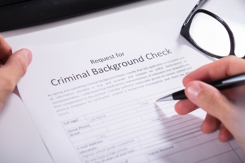 A close-up of a hand holding a pen, filling out a "Request for Criminal Background Check" form. The form includes fields for personal information. A pair of glasses and part of a keyboard are visible in the background, perhaps hinting at efforts to remove name from Radaris or similar databases.