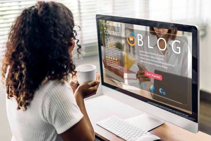 A person with curly hair sits at a desk, holding a mug and looking at a computer screen displaying a blog webpage. The desk has a keyboard and a mouse. The screen shows the word "BLOG" in large letters, alongside text asking, "What is a guest post?" accompanied by icons.