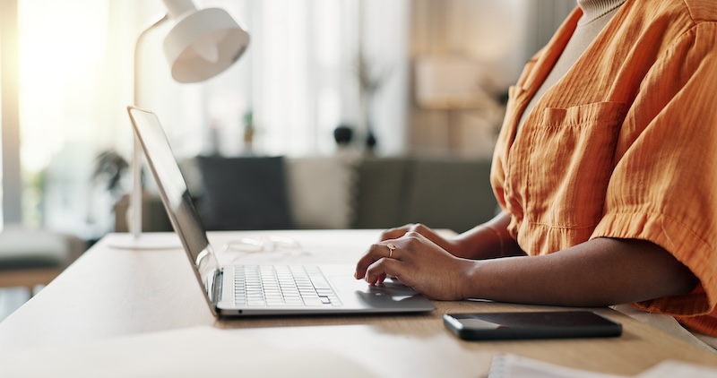 A person in an orange shirt works on a laptop at a desk in a well-lit room. A smartphone and some papers are placed beside the laptop, perhaps researching "what is a guest post." A white desk lamp is visible in the background, creating a cozy and casual setting.