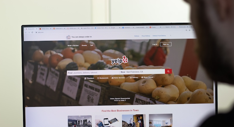 A person is looking at a computer screen displaying the Yelp homepage, exploring business reviews. The screen shows a search bar and images of fresh produce in the background, while various tabs are open at the top of the browser.